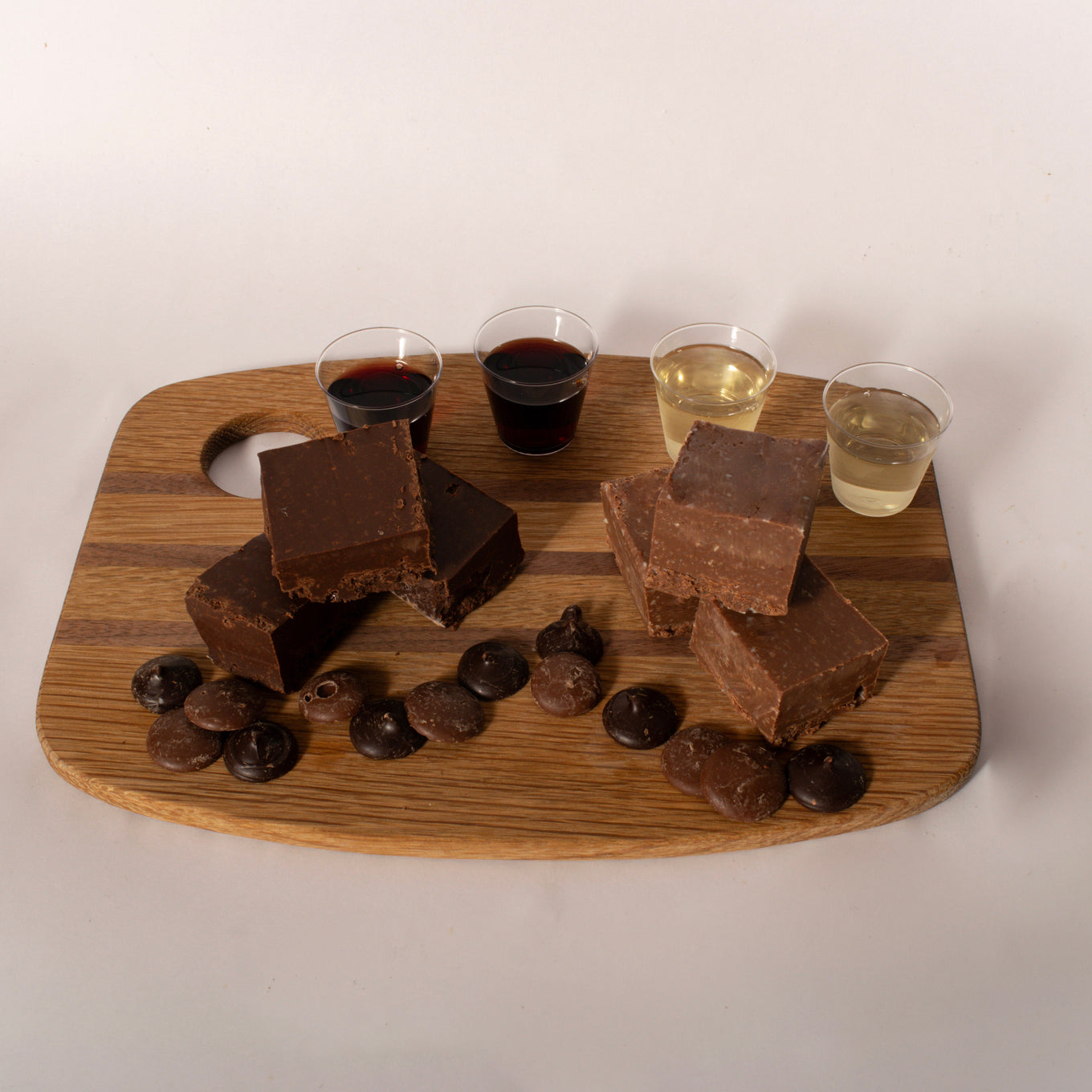 A wooden serving board with chocolate fudge bars, chocolate chips, and four small cups of different colored beverages, photographed against a plain off-white background.