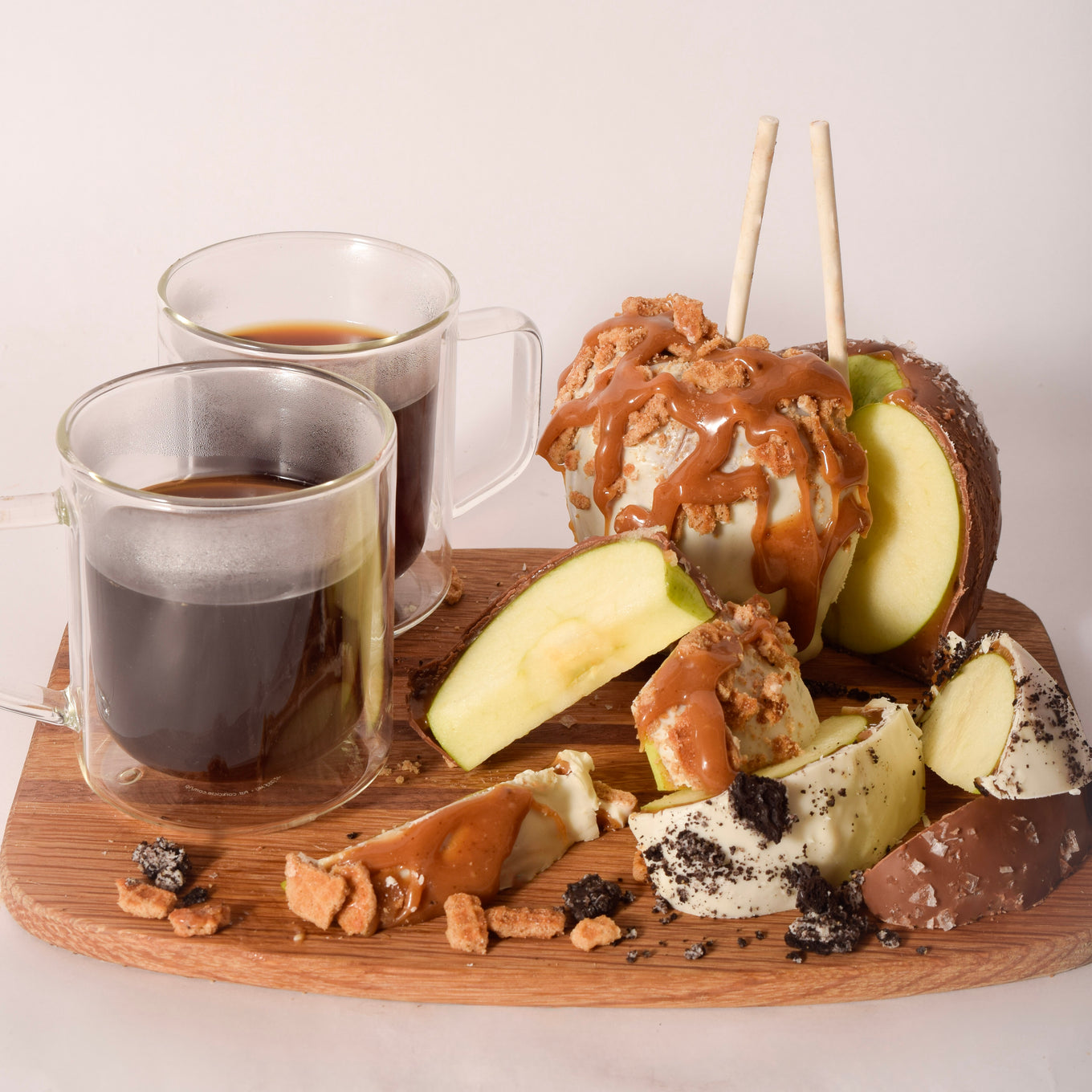 Two chocolate-dipped caramel apples on sticks, one topped with white chocolate, caramel drizzle, and streusel crumble, sliced open on a wood board with white and dark chocolate wedges, cookie crumble, and two glass mugs of coffee.
