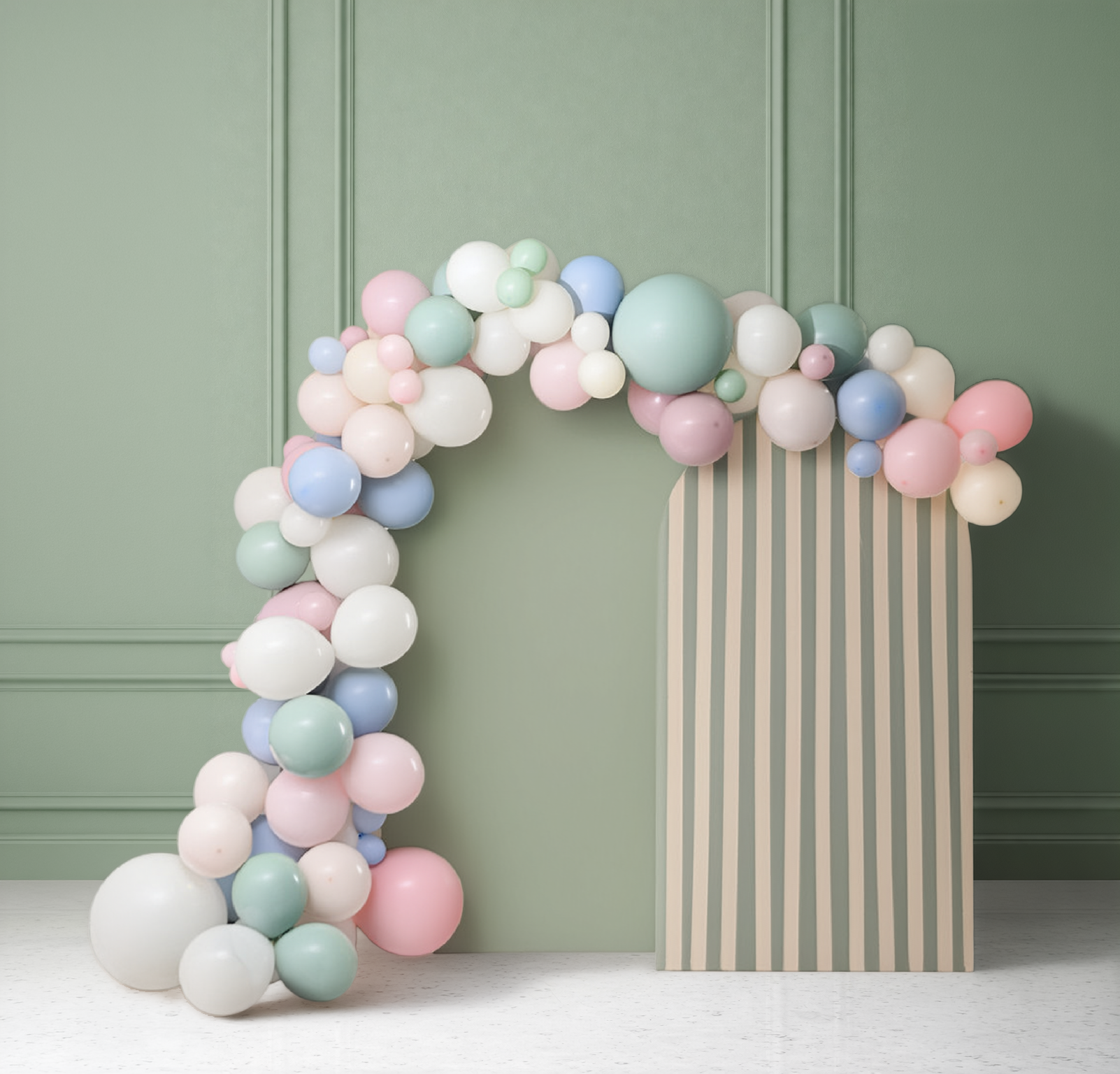 Pastel balloon garland arch in sage green, blush pink, dusty blue, ivory, and mint, curving beside a striped green-and-cream backdrop panel, set against a sage green paneled wall.