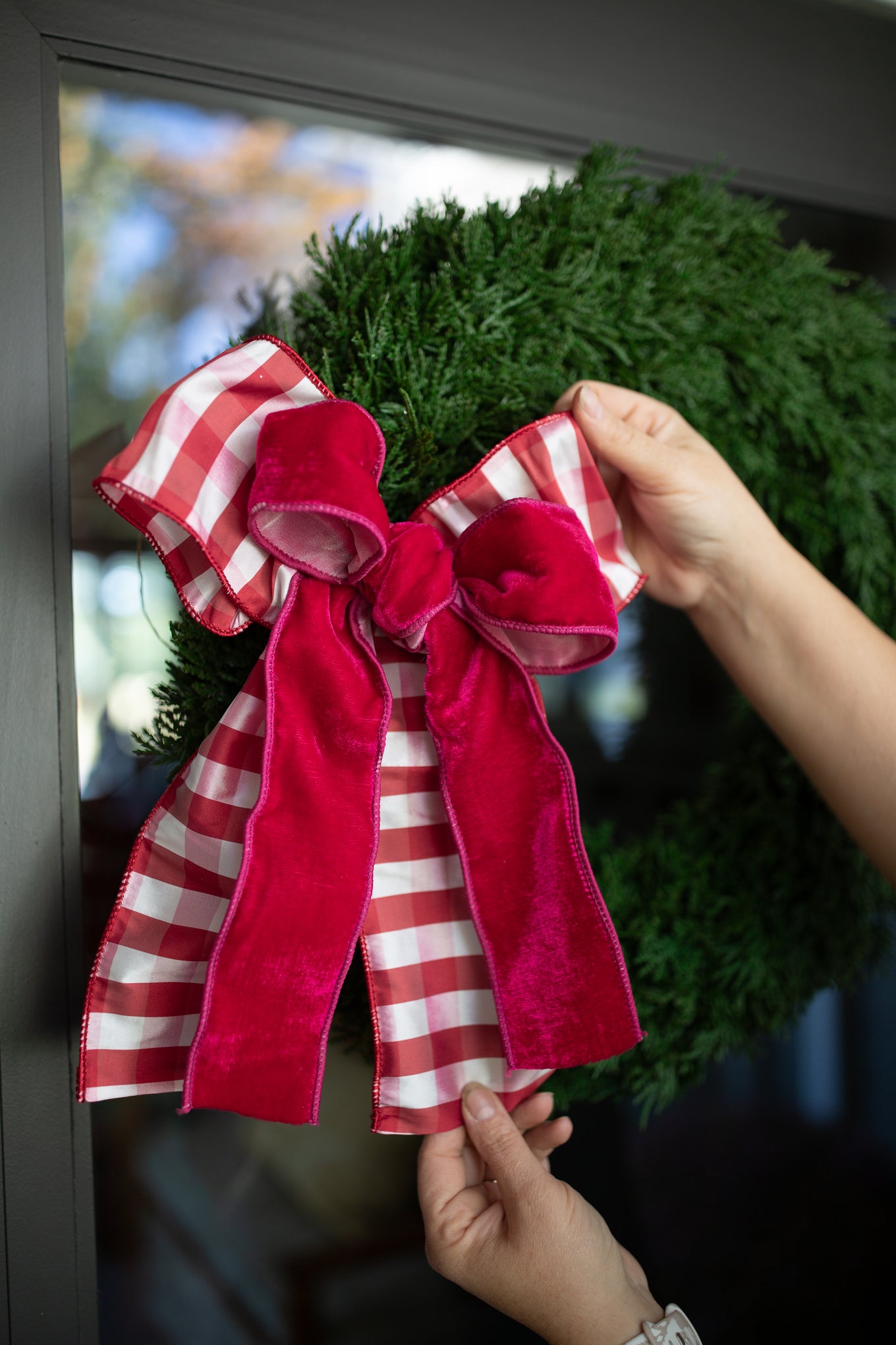 Hands adjusting a red velvet and red-and-white gingham bow on a fresh evergreen wreath.