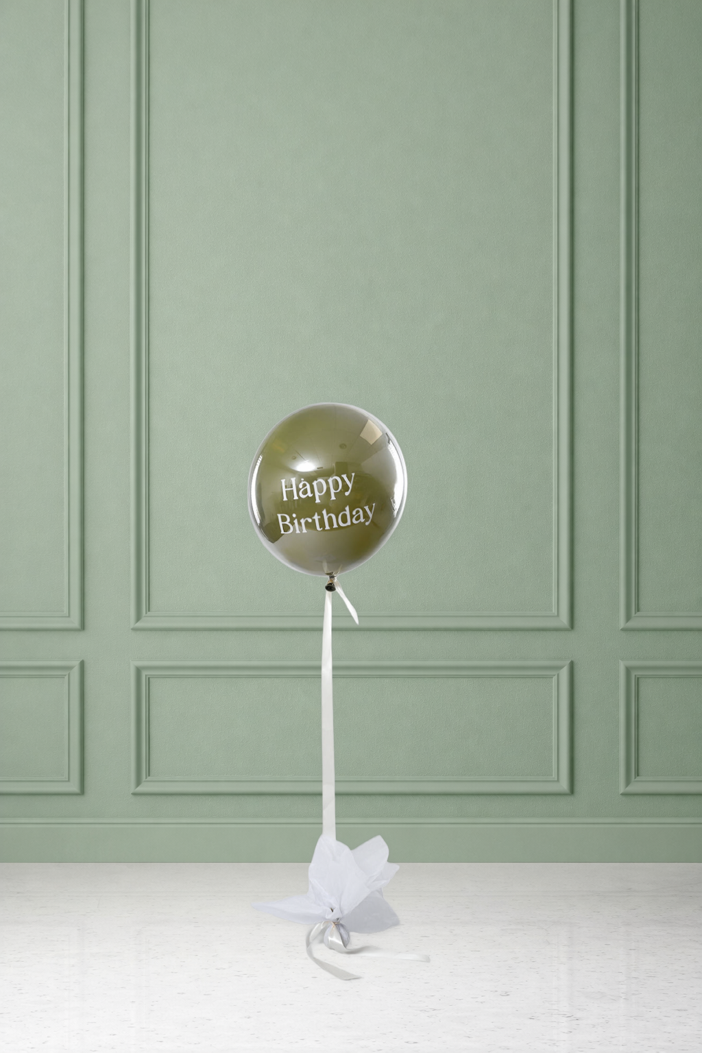 Single olive-green orbz balloon with 'Happy Birthday' printed in white text, tied with white ribbon and tissue paper to a weight, set against a sage green paneled wall.