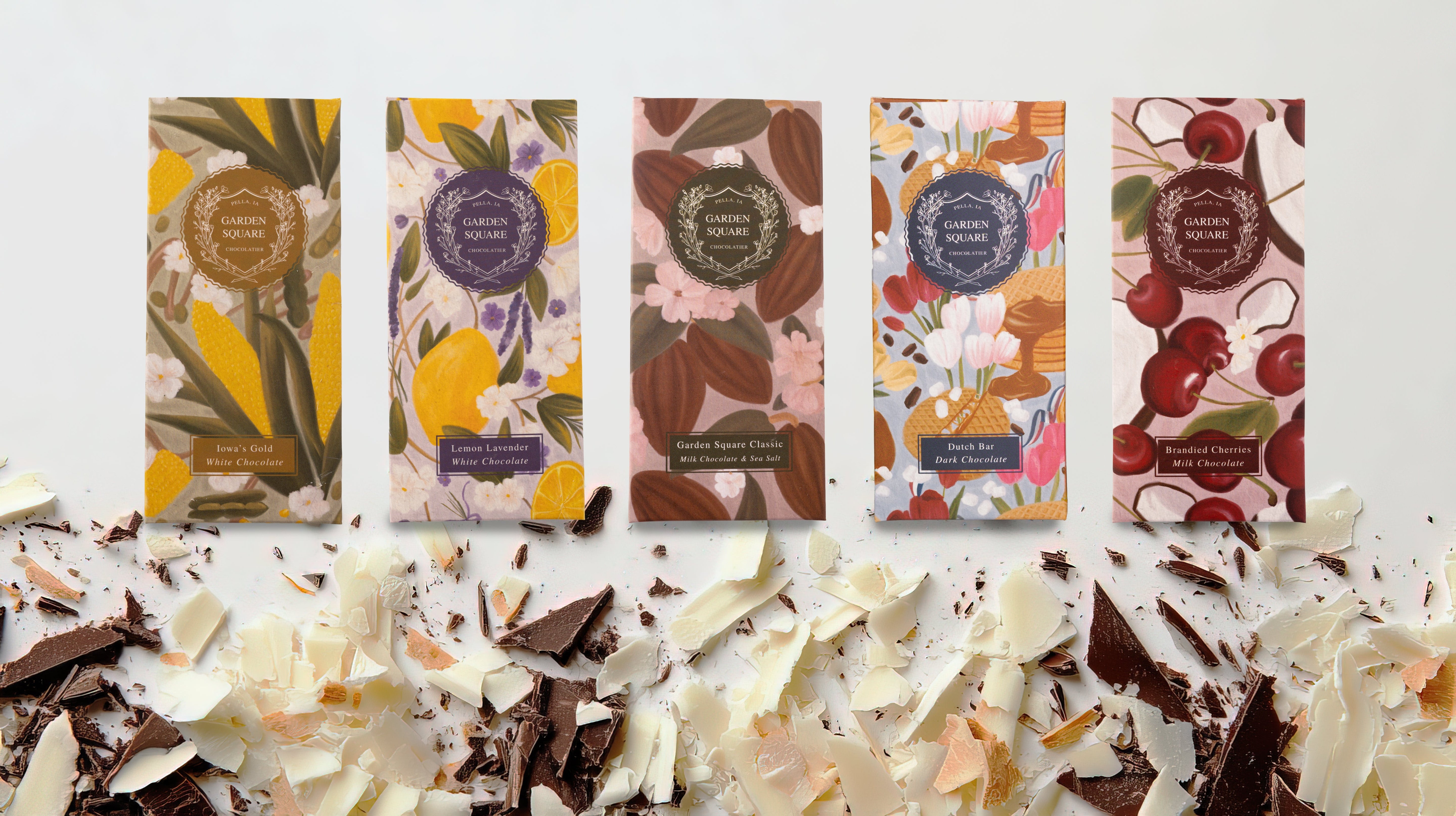 Five Garden Square Chocolatier bars—Iowa's Gold, Lemon Lavender, Classic, Dutch Bar, and Brandied Cherries—stand above a pile of white and dark chocolate shavings.