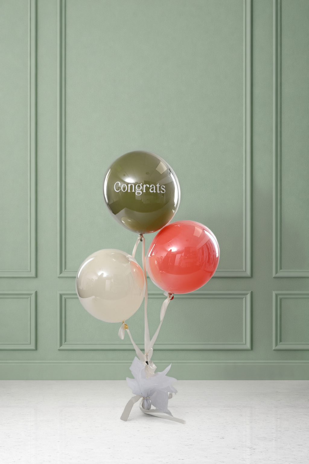Cluster of three orbz balloons — olive green printed with 'Congrats,' cream, and coral — tied with white ribbon and tissue paper to a weight, set against a sage green paneled wall.