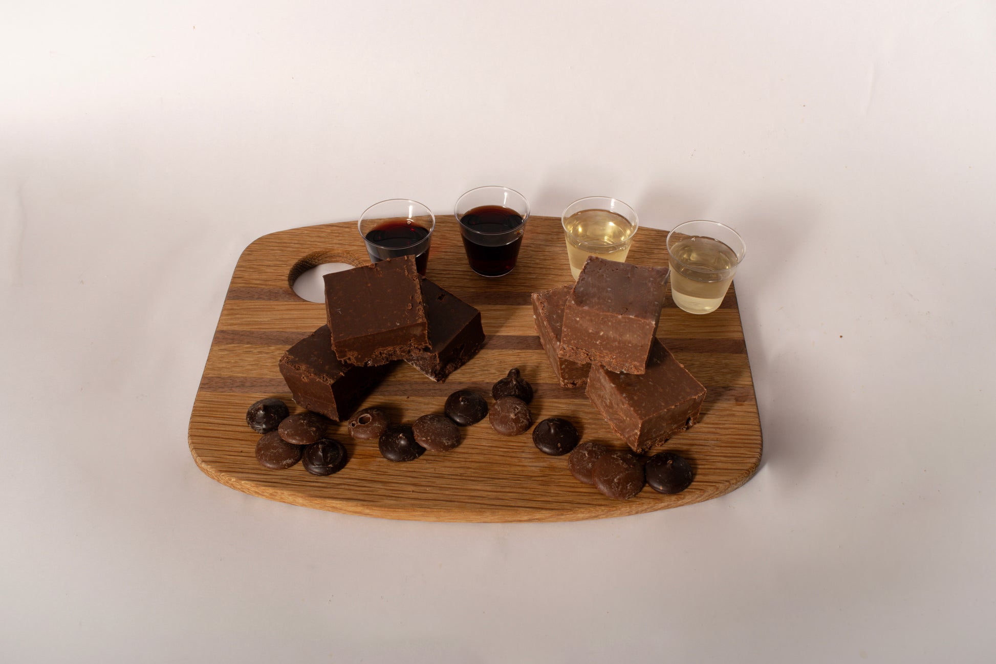 A wooden serving board with chocolate fudge bars, chocolate chips, and four small cups of different colored beverages, photographed against a plain off-white background.
