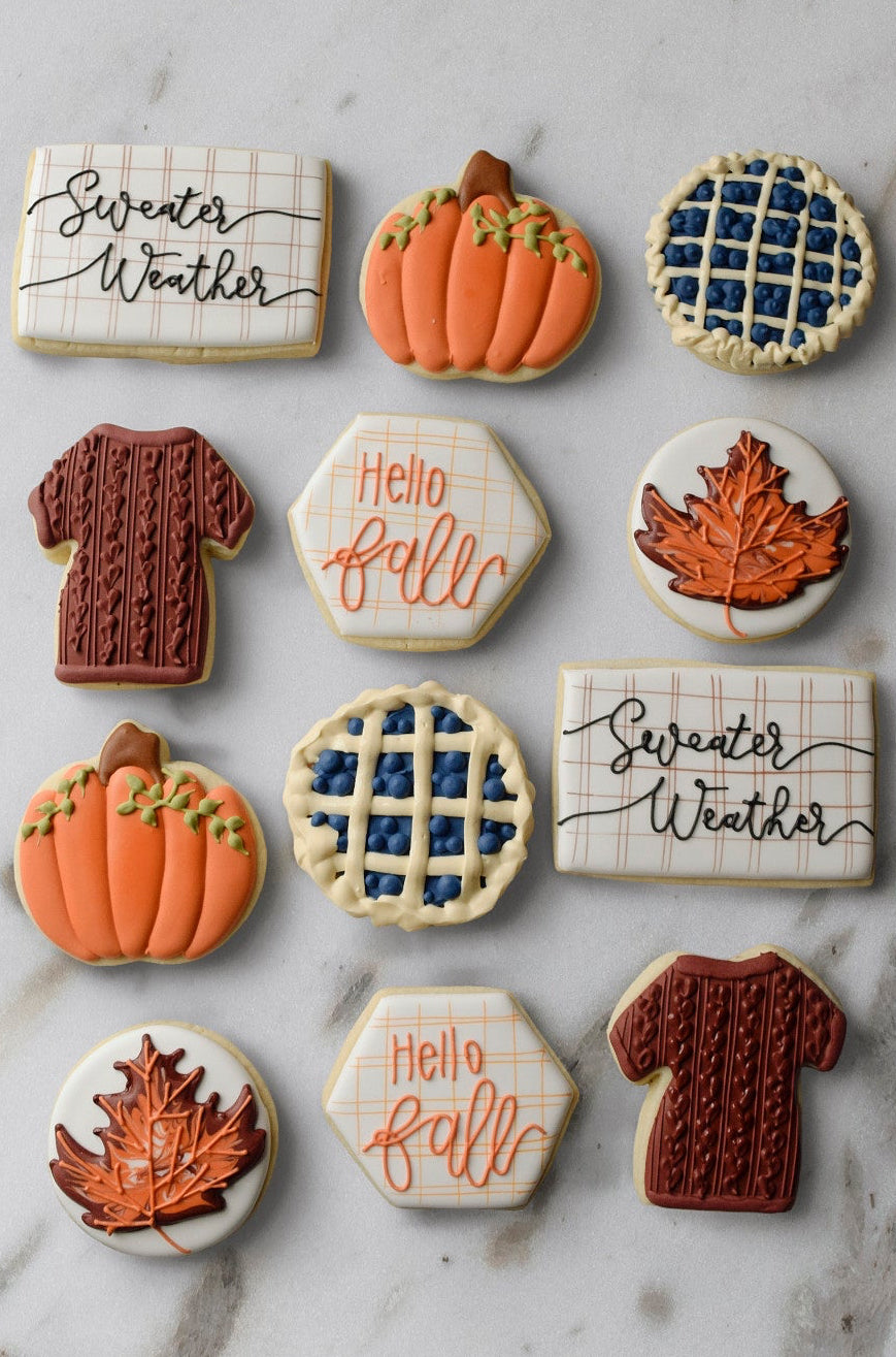 Hand-decorated fall cookies including "Sweater Weather" plaid rectangles, orange pumpkins, blueberry lattice pies, brown cable-knit sweaters, "Hello fall" plaid hexagons, and orange maple leaves on white marble, Pella, Iowa