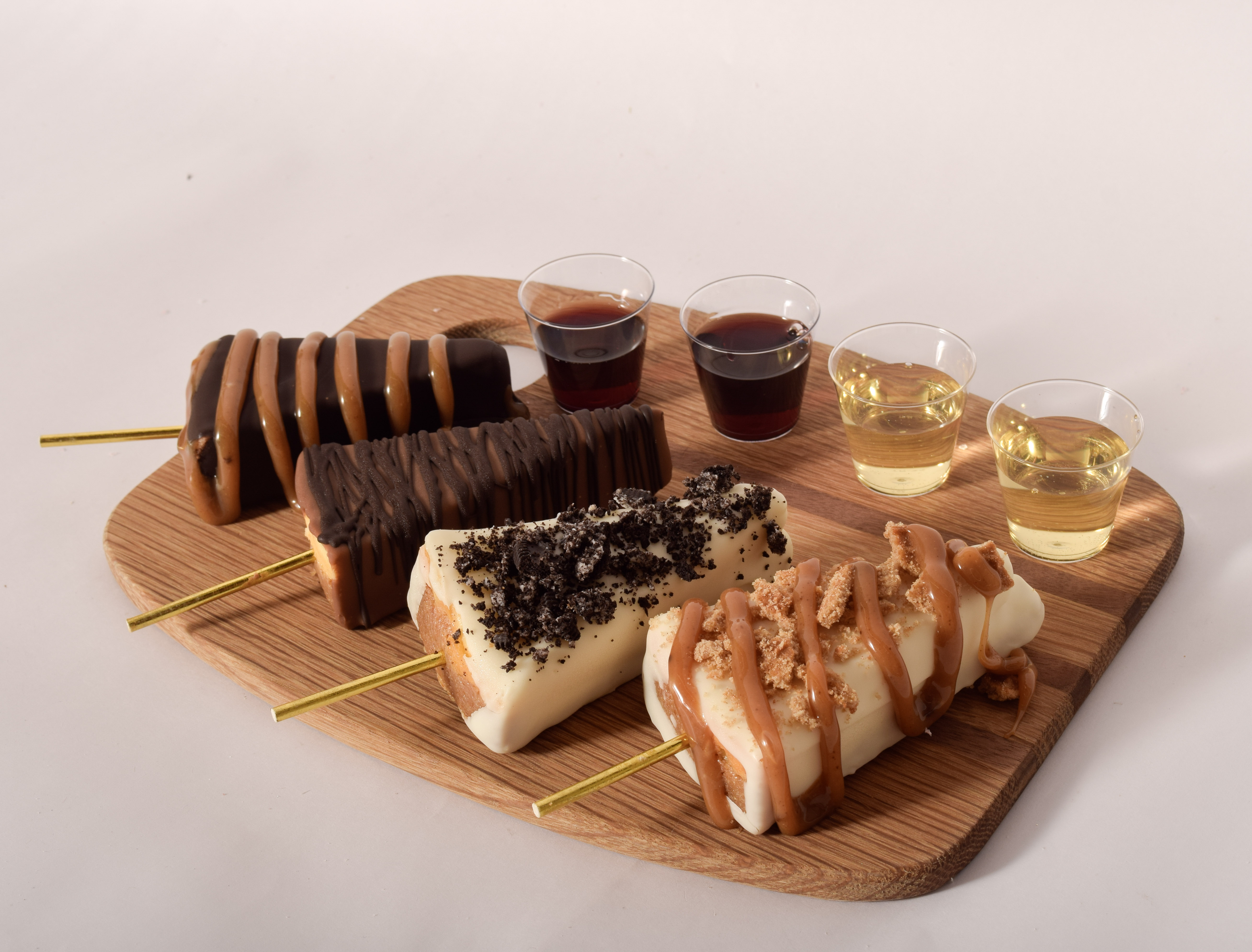 Four cheesecake pops on gold sticks — chocolate and caramel drizzle, chocolate striped, white chocolate with cookie crumble, and white chocolate with caramel and brown sugar crumble — arranged on a wood board next to four tasting glasses of red and white wine.
