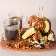 Two chocolate-dipped caramel apples on sticks, one topped with white chocolate, caramel drizzle, and streusel crumble, sliced open on a wood board with white and dark chocolate wedges, cookie crumble, and two glass mugs of coffee.