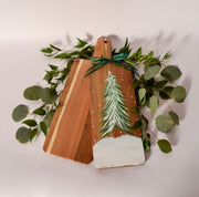 Two wooden cutting boards with a hand-painted Christmas tree design, wrapped in a green ribbon and surrounded by eucalyptus leaves on a light background.