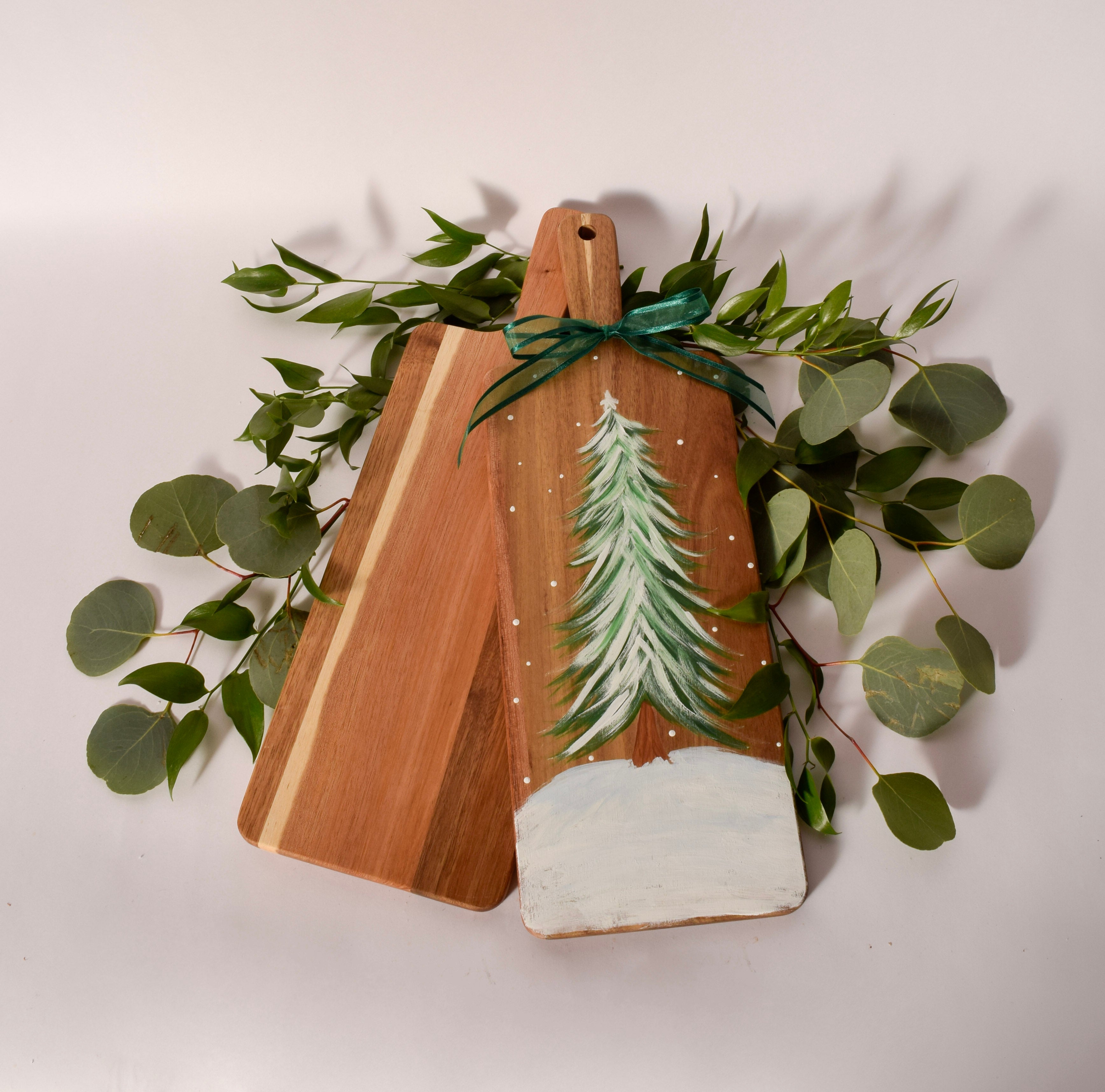 Two wooden cutting boards with a hand-painted Christmas tree design, wrapped in a green ribbon and surrounded by eucalyptus leaves on a light background.