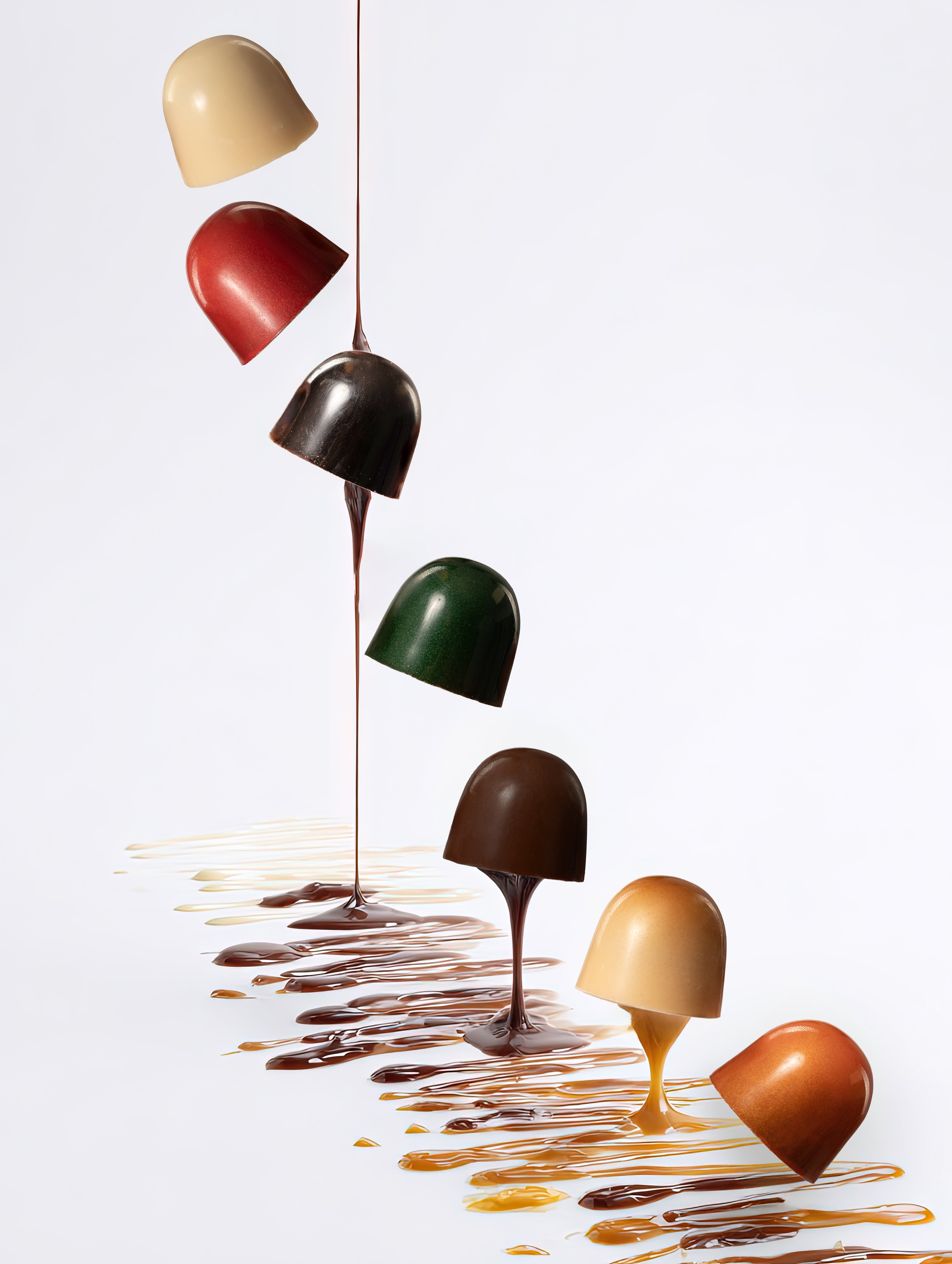 Six dome-shaped chocolate truffles in assorted colors are suspended vertically above a glossy white surface, with melted chocolate dripping from each and pooling below in a decorative pattern.