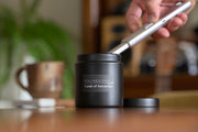 Holland Scents Canals of Amsterdam soy candle being lit on a wooden table with a coffee mug and plant, Pella, Iowa