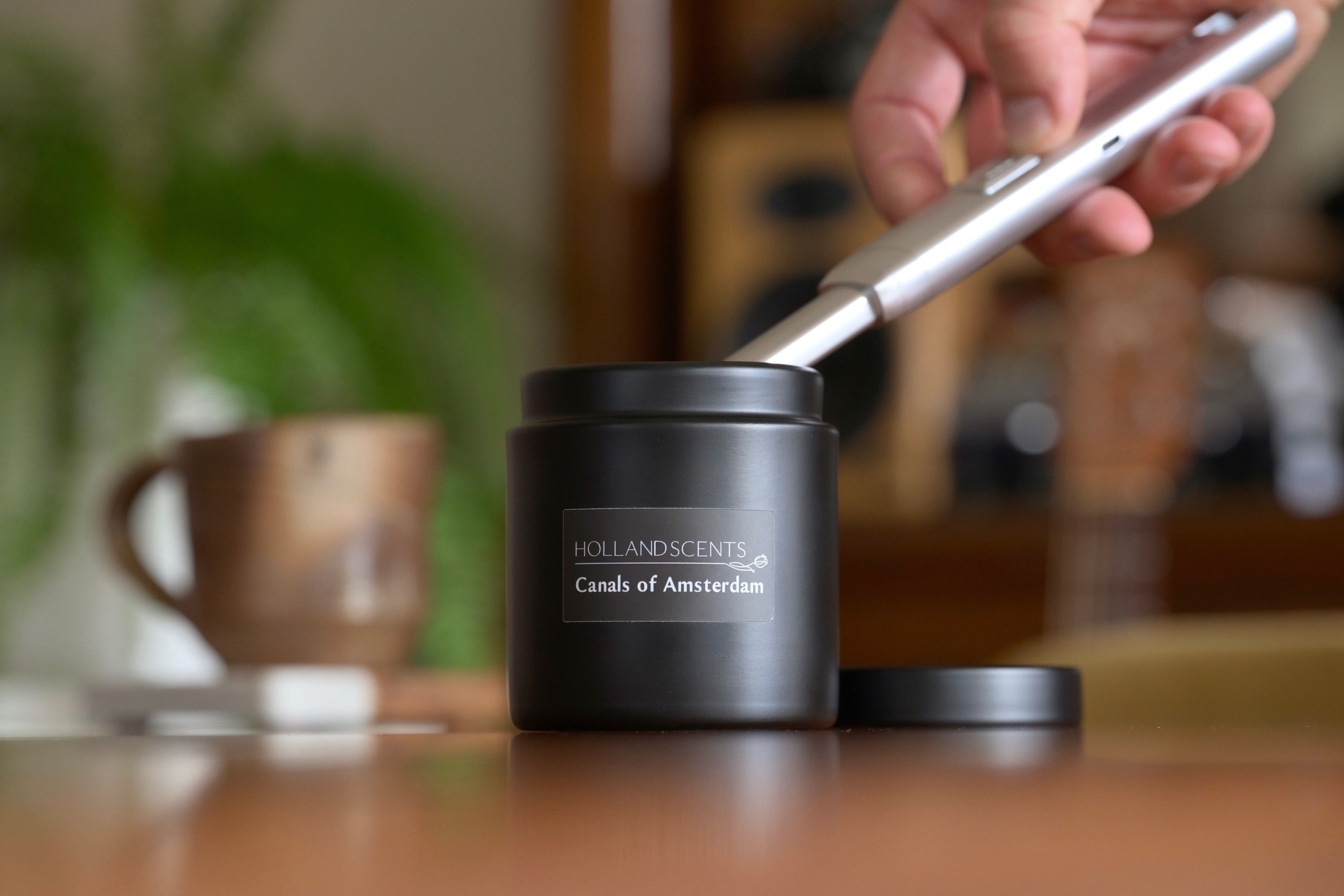 Holland Scents Canals of Amsterdam soy candle being lit on a wooden table with a coffee mug and plant, Pella, Iowa