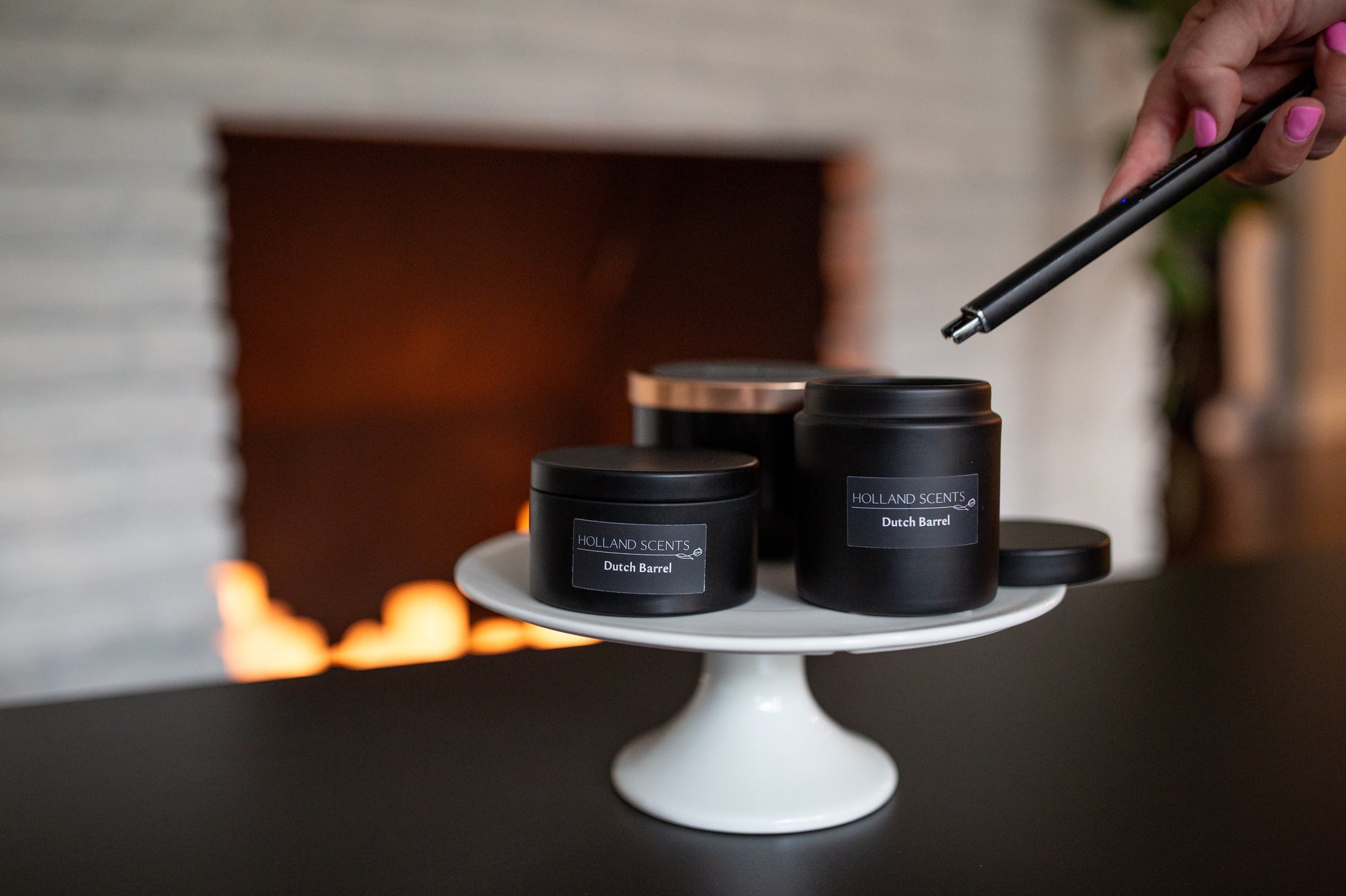 Three sizes of Holland Scents Dutch Barrel soy candles on a white cake stand near a lit fireplace, Pella, Iowa