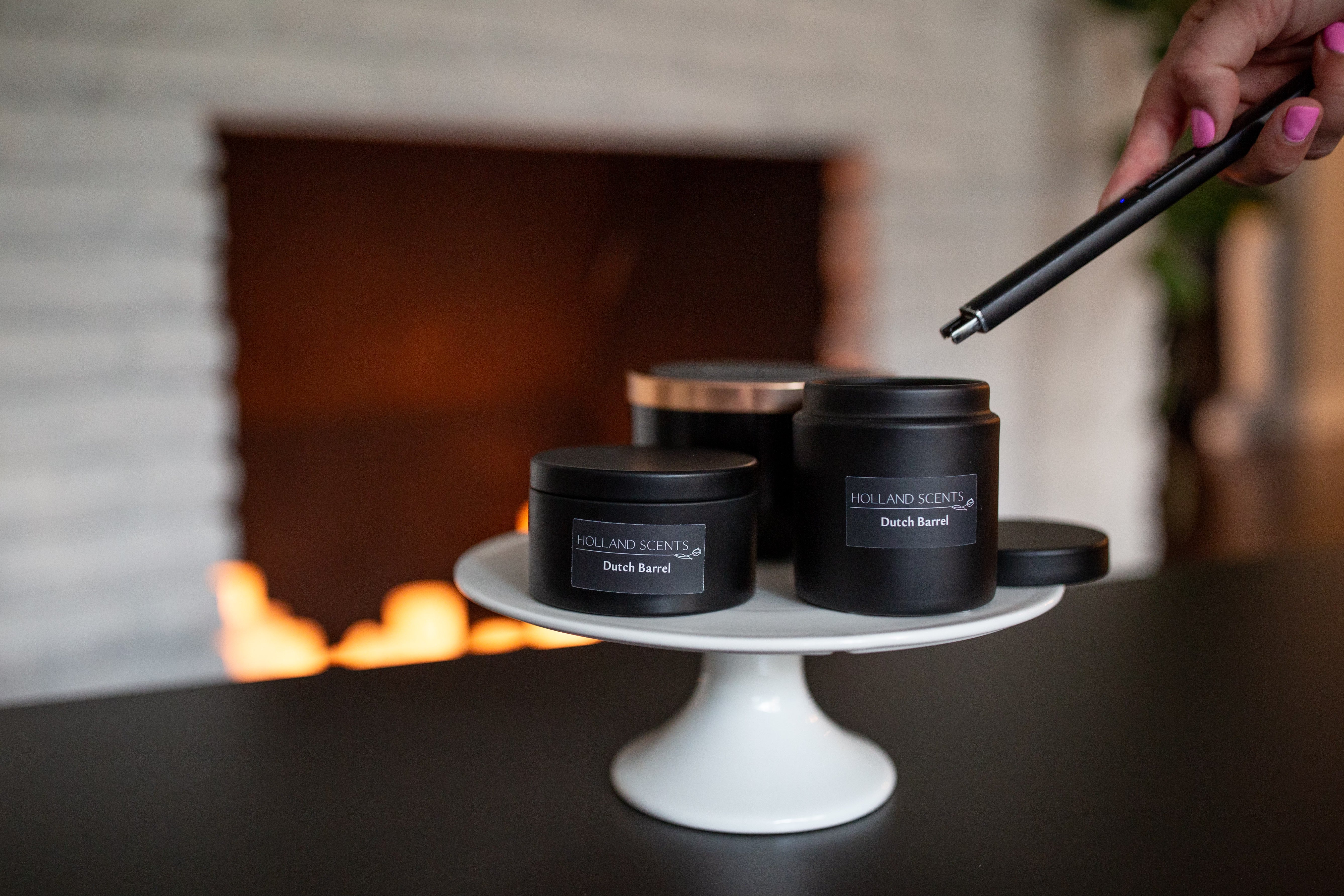 Three sizes of Holland Scents Dutch Barrel soy candles on a white cake stand near a lit fireplace, Pella, Iowa