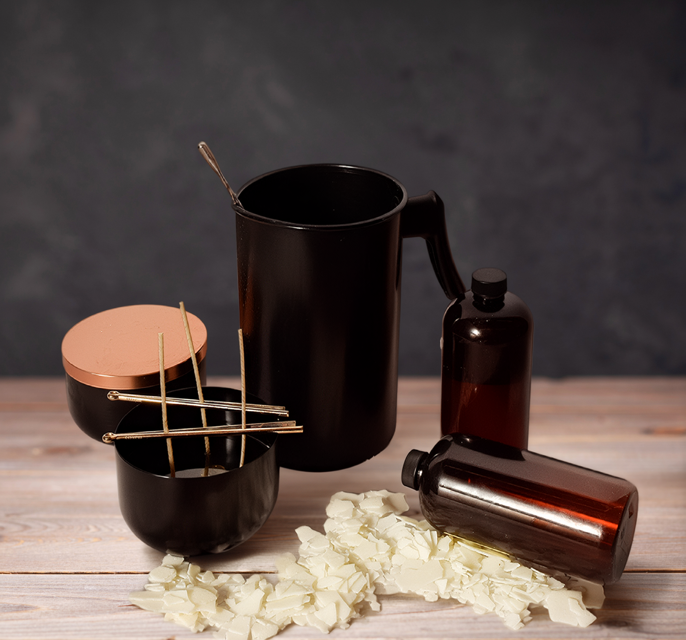 Candle-making supplies including a black wax pouring pitcher, black candle vessels with copper lids and wick centering tools, amber fragrance oil bottles, and soy wax flakes on a wood table.