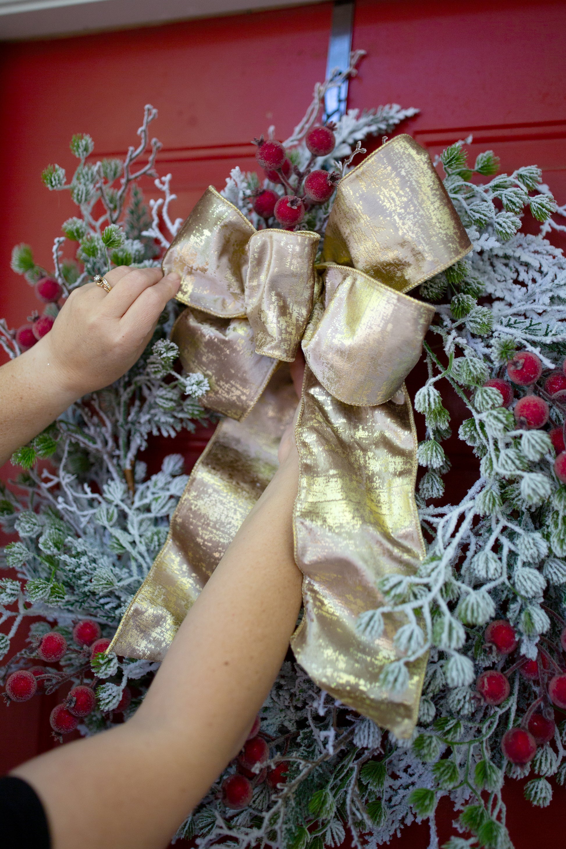 Hands adjusting a champagne gold metallic bow on a flocked evergreen wreath with red berries, hung on a red door.