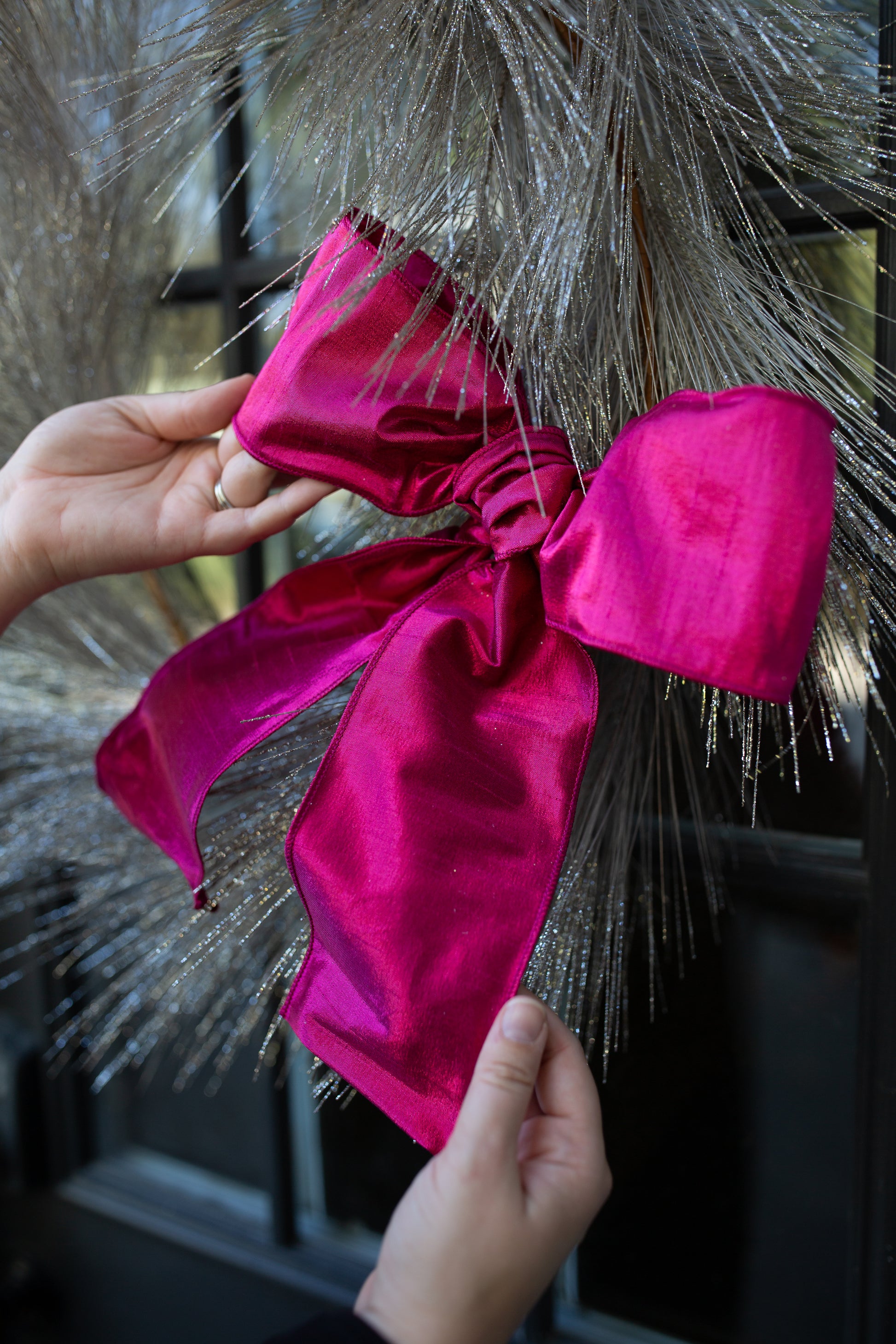 Hands tying a fuchsia pink bow into a silvery, glittered tinsel-style wreath or tree.