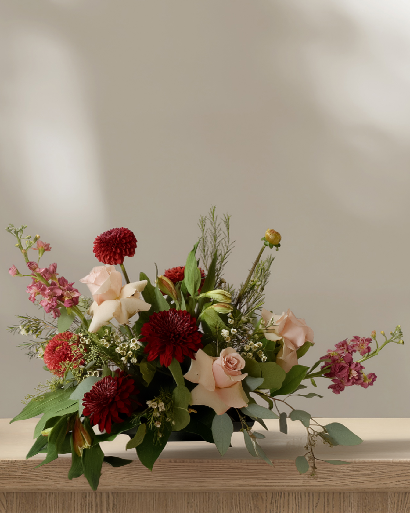Low, wide Thanksgiving floral centerpiece with burgundy dahlias, blush roses, cream lilies, pink stock, and eucalyptus greenery in a low black vase, styled on a wood table.