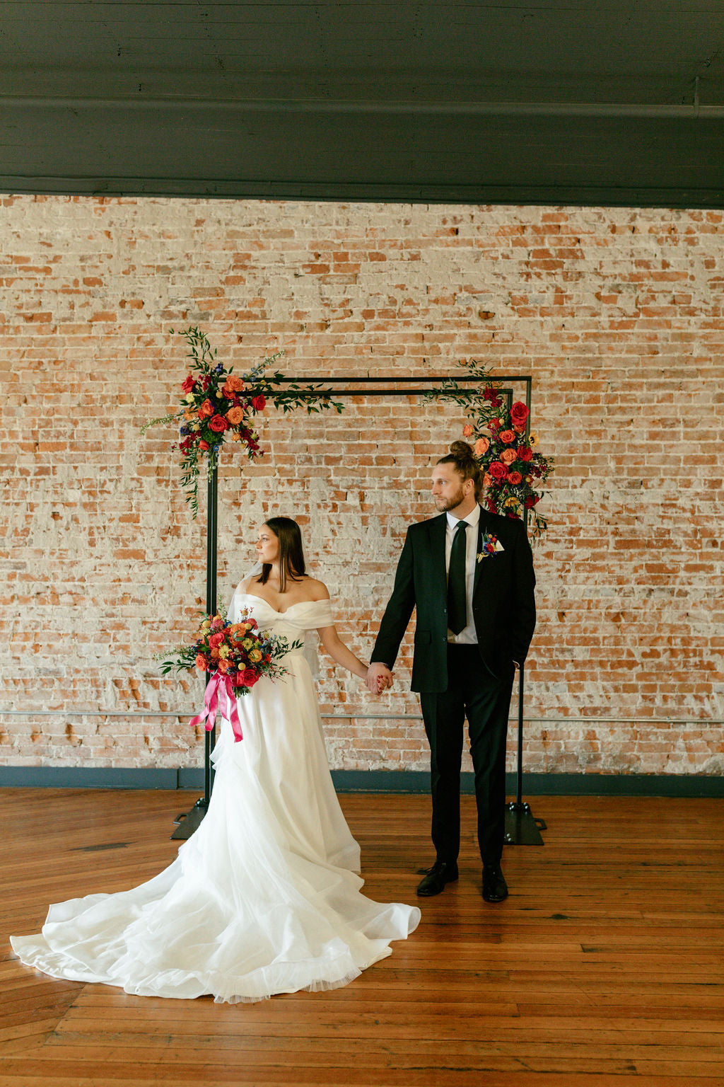 A bride in a white off-the-shoulder wedding gown and a groom in a black suit holding hands, standing in front of a rustic brick wall backdrop with a floral arch.